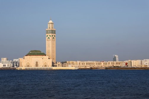 A prominent mosque stands near the waterfront, characterized by its large minaret adorned with intricate designs. The structure features a blend of traditional Islamic architecture with modern city buildings visible in the background under a clear blue sky.