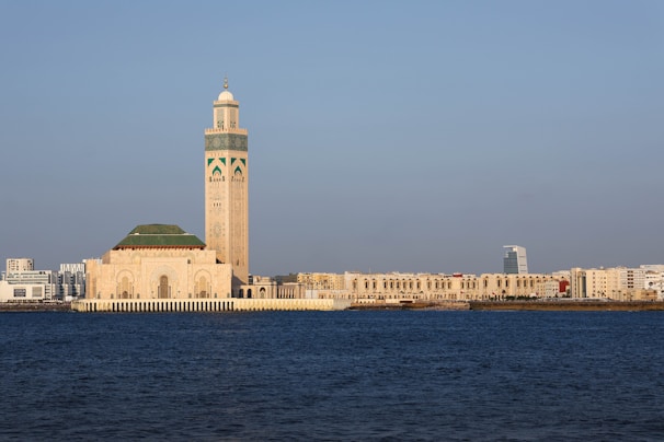 A prominent mosque stands near the waterfront, characterized by its large minaret adorned with intricate designs. The structure features a blend of traditional Islamic architecture with modern city buildings visible in the background under a clear blue sky.