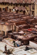 A traditional tannery with multiple circular vats filled with different colored liquids, surrounded by an old building with drying hides hanging along the walls. Workers are seen tending to the vats and processing hides using long sticks and tools.