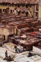 A traditional tannery with multiple circular vats filled with different colored liquids, surrounded by an old building with drying hides hanging along the walls. Workers are seen tending to the vats and processing hides using long sticks and tools.