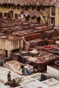 A traditional tannery with multiple circular vats filled with different colored liquids, surrounded by an old building with drying hides hanging along the walls. Workers are seen tending to the vats and processing hides using long sticks and tools.
