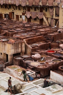 A traditional tannery with multiple circular vats filled with different colored liquids, surrounded by an old building with drying hides hanging along the walls. Workers are seen tending to the vats and processing hides using long sticks and tools.