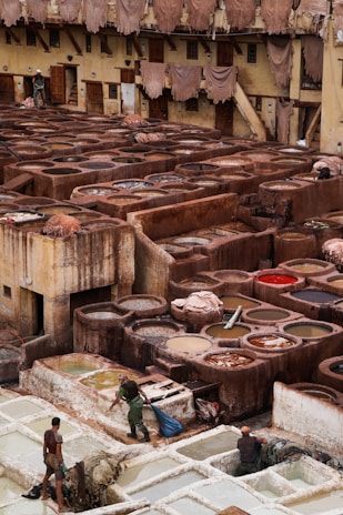 A traditional tannery with multiple circular vats filled with different colored liquids, surrounded by an old building with drying hides hanging along the walls. Workers are seen tending to the vats and processing hides using long sticks and tools.