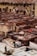 A traditional tannery with multiple circular vats filled with different colored liquids, surrounded by an old building with drying hides hanging along the walls. Workers are seen tending to the vats and processing hides using long sticks and tools.