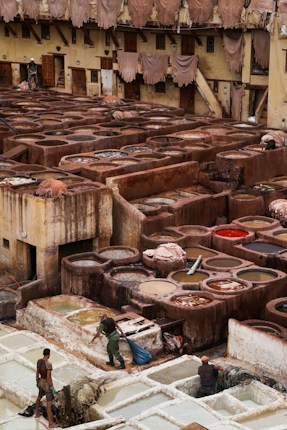 A traditional tannery with multiple circular vats filled with different colored liquids, surrounded by an old building with drying hides hanging along the walls. Workers are seen tending to the vats and processing hides using long sticks and tools.