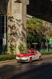 A red and white taxi is driving on a sunny street under a large concrete structure. There are green plants and trees casting shadows on the ground. The background shows a building with large glass windows and green signage.