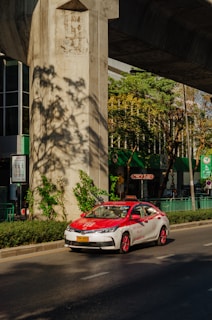 A red and white taxi is driving on a sunny street under a large concrete structure. There are green plants and trees casting shadows on the ground. The background shows a building with large glass windows and green signage.