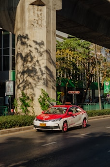 A red and white taxi is driving on a sunny street under a large concrete structure. There are green plants and trees casting shadows on the ground. The background shows a building with large glass windows and green signage.