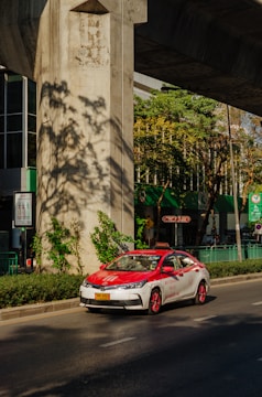 A red and white taxi is driving on a sunny street under a large concrete structure. There are green plants and trees casting shadows on the ground. The background shows a building with large glass windows and green signage.