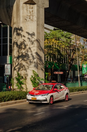 A red and white taxi is driving on a sunny street under a large concrete structure. There are green plants and trees casting shadows on the ground. The background shows a building with large glass windows and green signage.