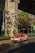 A red and white taxi is driving on a sunny street under a large concrete structure. There are green plants and trees casting shadows on the ground. The background shows a building with large glass windows and green signage.