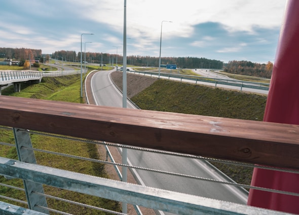 A view from a bridge overlooking a multi-lane highway with several overpasses. The road is surrounded by grassy embankments and bordered by metal railings. The sky is mostly cloudy with patches of blue visible, and there's a forested area in the background with autumn foliage.