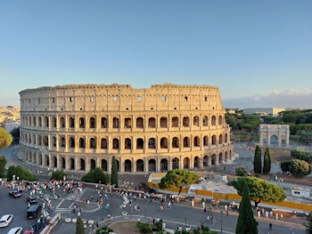 The image features an ancient Roman amphitheater with its distinctive arched structure surrounded by a throng of tourists and visitors. The scene is set under a clear blue sky, with surrounding greenery and a large historical triumphal arch in the background.