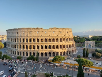 The image features an ancient Roman amphitheater with its distinctive arched structure surrounded by a throng of tourists and visitors. The scene is set under a clear blue sky, with surrounding greenery and a large historical triumphal arch in the background.