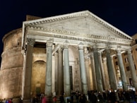 A grand ancient Roman building with tall Corinthian columns at the entrance and a large triangular pediment above. The structure is well-lit against a night sky, highlighting the weathered stone and intricate architectural details. The inscription 'M•AGRIPPA•L•F•COS•TERTIVM•FECIT' is visible on the pediment. People are gathered near the entrance, showing the scale of the structure.