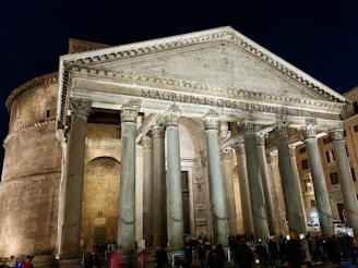 A grand ancient Roman building with tall Corinthian columns at the entrance and a large triangular pediment above. The structure is well-lit against a night sky, highlighting the weathered stone and intricate architectural details. The inscription 'M•AGRIPPA•L•F•COS•TERTIVM•FECIT' is visible on the pediment. People are gathered near the entrance, showing the scale of the structure.