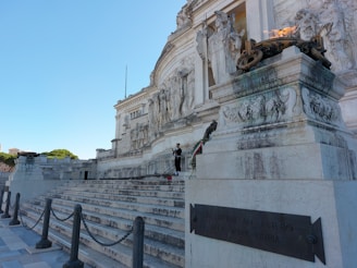 A large marble monument with intricate carvings and statues, featuring stairs leading up to the structure. An eternal flame burns in a large bowl on the right. A person stands near the monument, adding a human element to the scene. The monument has classical architectural features and relief sculptures.
