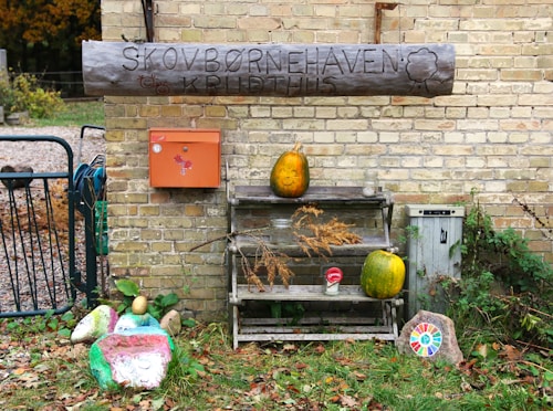 A wooden sign with engraved text is mounted on a brick wall, accompanied by an orange mailbox. Below, a wooden shelf holds pumpkins, with one carved and painted with a face. Various objects, including a painted stone with a colorful sun design, are placed around the setup, amidst some greenery and scattered leaves.
