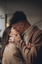 A couple is intimately embracing and sharing a kiss. They are both wearing brown coats, and their expressions suggest affection and closeness. The setting appears to be indoors with soft lighting, giving a warm atmosphere.