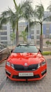 A vibrant red car is parked on a cobblestone surface, flanked by palm trees. The vehicle's design is sleek and sporty, bearing a distinctive front grille with the logo in the center. The building in the background features large windows, painted in grey and blue tones. A black car is partially visible on the left side.