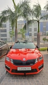 A vibrant red car is parked on a cobblestone surface, flanked by palm trees. The vehicle's design is sleek and sporty, bearing a distinctive front grille with the logo in the center. The building in the background features large windows, painted in grey and blue tones. A black car is partially visible on the left side.