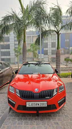 A vibrant red car is parked on a cobblestone surface, flanked by palm trees. The vehicle's design is sleek and sporty, bearing a distinctive front grille with the logo in the center. The building in the background features large windows, painted in grey and blue tones. A black car is partially visible on the left side.