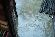 A wet patio area with several puddles reflecting raindrop impact, outlined by a brick wall and a welcome mat with an open door. The surface is shiny and slick due to the rain.