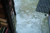A wet patio area with several puddles reflecting raindrop impact, outlined by a brick wall and a welcome mat with an open door. The surface is shiny and slick due to the rain.