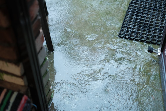 A wet patio area with several puddles reflecting raindrop impact, outlined by a brick wall and a welcome mat with an open door. The surface is shiny and slick due to the rain.