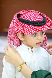 A young boy wearing a traditional red and white keffiyeh with a black agal. His white thobe is buttoned up. An adult's hands adjust the headscarf, one of which is wearing a gold watch.