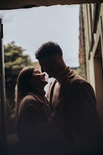 A couple stands close together, sharing an intimate moment. The scene is softly lit, creating a romantic and warm atmosphere. They are silhouetted against a soft background of trees and architecture, suggesting an outdoor setting.