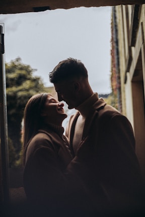 A couple stands close together, sharing an intimate moment. The scene is softly lit, creating a romantic and warm atmosphere. They are silhouetted against a soft background of trees and architecture, suggesting an outdoor setting.