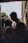 A couple stands close together, sharing an intimate moment. The scene is softly lit, creating a romantic and warm atmosphere. They are silhouetted against a soft background of trees and architecture, suggesting an outdoor setting.