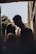 A couple stands close together, sharing an intimate moment. The scene is softly lit, creating a romantic and warm atmosphere. They are silhouetted against a soft background of trees and architecture, suggesting an outdoor setting.