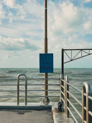 A metal railing lines the edge of a concrete pier, extending into a body of water. A vertical pole supports a blue sign with the words 'Sea You Soon' printed in a bold font. The sky is partly cloudy, and the sea appears slightly choppy, with gentle waves visible.