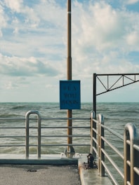 A metal railing lines the edge of a concrete pier, extending into a body of water. A vertical pole supports a blue sign with the words 'Sea You Soon' printed in a bold font. The sky is partly cloudy, and the sea appears slightly choppy, with gentle waves visible.