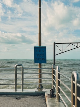 A metal railing lines the edge of a concrete pier, extending into a body of water. A vertical pole supports a blue sign with the words 'Sea You Soon' printed in a bold font. The sky is partly cloudy, and the sea appears slightly choppy, with gentle waves visible.