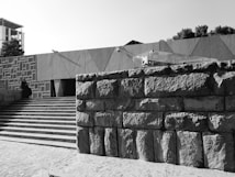 A black and white photograph depicts a modern architectural setting with geometric patterns on the walls. A person dressed in dark clothing sits to the left on a staircase that leads up to a minimalist structure. Large stone blocks form a wall in the foreground, while sparse trees are visible in the background.