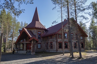 A rustic wooden building with a steep, pointed roof is set amidst a forest of tall trees. The building features several windows with red frames and has a unique structure with a small entrance porch. Two people can be seen near the entrance, giving a sense of scale. Sunlight filters through the trees, casting shadows on the ground.