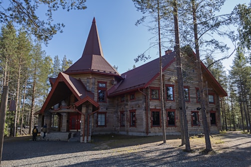 A rustic wooden building with a steep, pointed roof is set amidst a forest of tall trees. The building features several windows with red frames and has a unique structure with a small entrance porch. Two people can be seen near the entrance, giving a sense of scale. Sunlight filters through the trees, casting shadows on the ground.