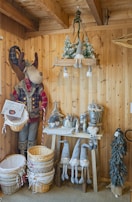 A cozy room with wooden walls is decorated with a large plush reindeer figure wearing a plaid jacket and furry boots. It stands next to a stack of wicker baskets. Nearby, a small table holds various decorative items such as metal cans and a line of gnome figurines with long legs and pointed hats. Above, a hanging wooden shelf displays snowy tree decorations. The overall theme is rustic and festive.