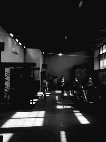 A dimly lit room features strong contrasts of light and shadow, with several individuals standing in various positions. The space has high ceilings and large windows casting patterned shadows on the floor. The focus is on the interplay of light and dark, creating a dramatic atmosphere.