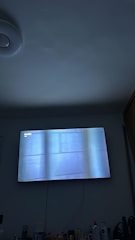 A dimly lit room with a television on the wall displaying a blue-tinted image. A variety of personal care products are visible on a surface below the TV. The ceiling has a circular light fixture, and a part of a door or cabinet is on the right side of the image.