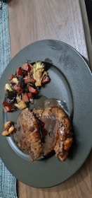 A serving of two slices of meat covered in a rich brown sauce is placed on a dark-colored plate. Accompanying the meat are roasted vegetables including broccoli and carrots, with some nuts visible. The plate sits on a wooden table next to a woven placemat.