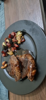 A serving of two slices of meat covered in a rich brown sauce is placed on a dark-colored plate. Accompanying the meat are roasted vegetables including broccoli and carrots, with some nuts visible. The plate sits on a wooden table next to a woven placemat.