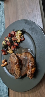 A serving of two slices of meat covered in a rich brown sauce is placed on a dark-colored plate. Accompanying the meat are roasted vegetables including broccoli and carrots, with some nuts visible. The plate sits on a wooden table next to a woven placemat.