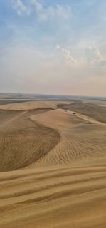 A vast desert landscape with rolling sand dunes under a partly cloudy sky. Multiple vehicles are seen driving across the sand, leaving tracks. The scene is expansive, evoking a sense of openness and adventure.