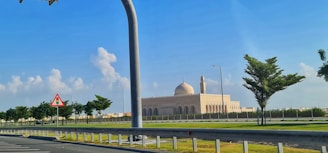 A large mosque with a prominent dome and a minaret is situated in a well-maintained area. Surrounding the mosque are neatly trimmed trees and a road in the foreground. A road sign and streetlights are visible, under a blue sky with a few clouds.
