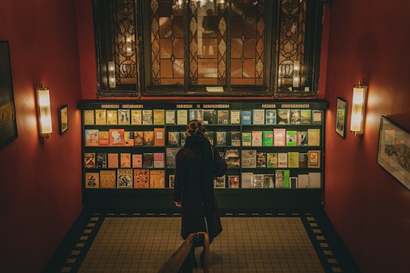A person stands in front of a wall of colorful books arranged on shelves, located in a dimly lit room with dark red walls and vintage light fixtures. The setting appears to be an intimate, cozy library or bookstore with a classic, slightly nostalgic atmosphere.