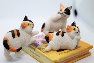 Three small, colorful cat figurines are positioned atop a closed book. The cats are animated in various playful poses. The book appears to be written in a language other than English, with some text visible on the cover. The background is minimal and out of focus, ensuring the primary focus remains on the figures and the book.